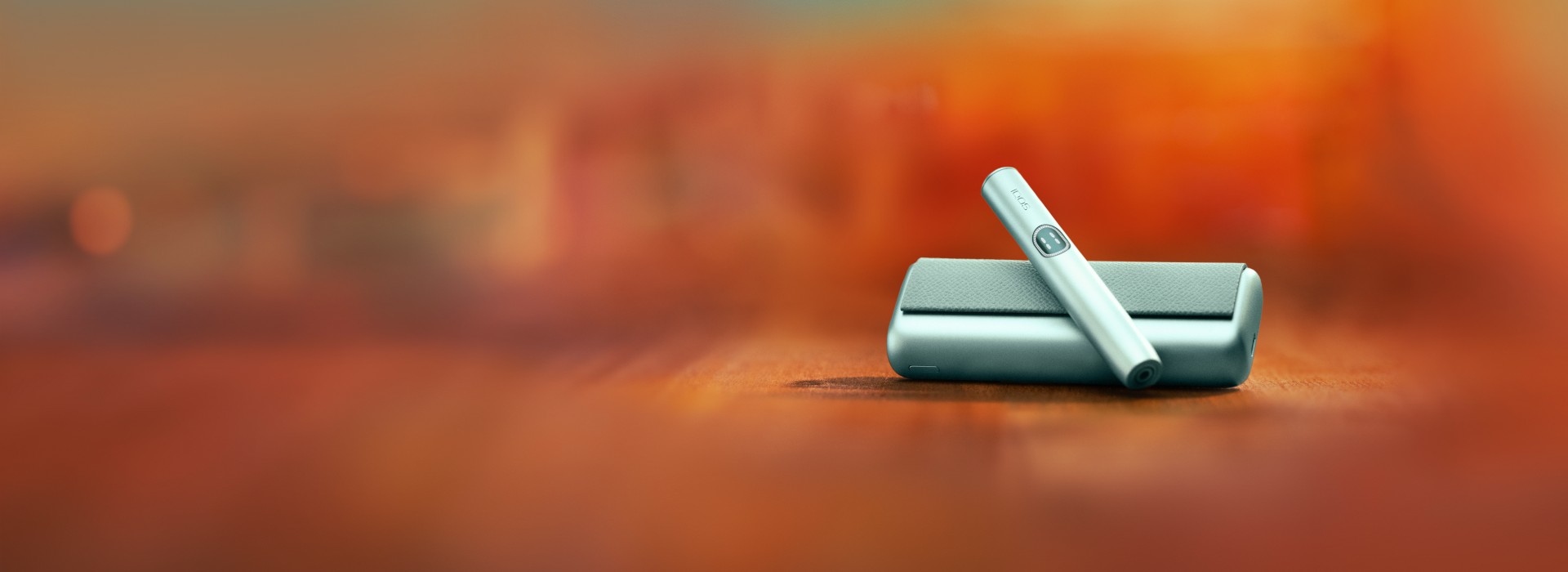 Close-up of an IQOS ILUMA i PRIME. The holder is resting on top of its pocket charger, set against a warm orange-red gradient background.